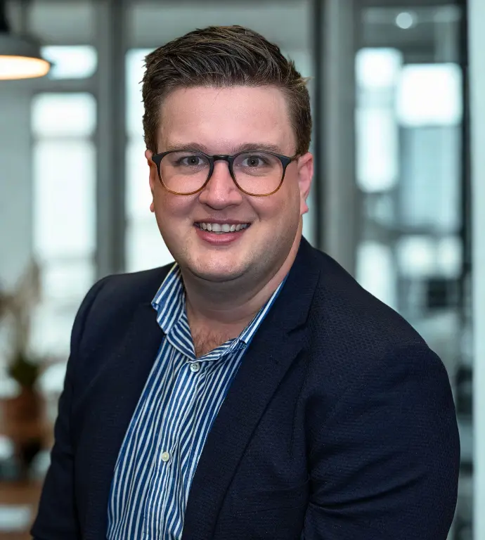Grant Peel, founder of Niique &amp; Co, smiling in a modern office setting. Wearing glasses, a navy blazer, and a blue-striped shirt, he exudes a confident, approachable presence.